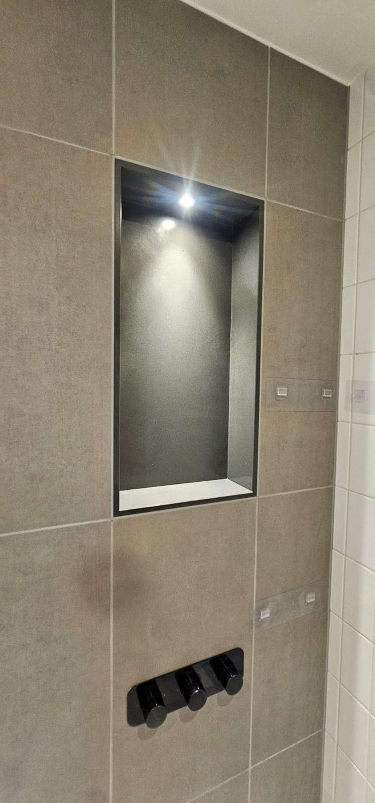 Recessed shower niche in a grey tiled wall with a spotlight and black shower controls