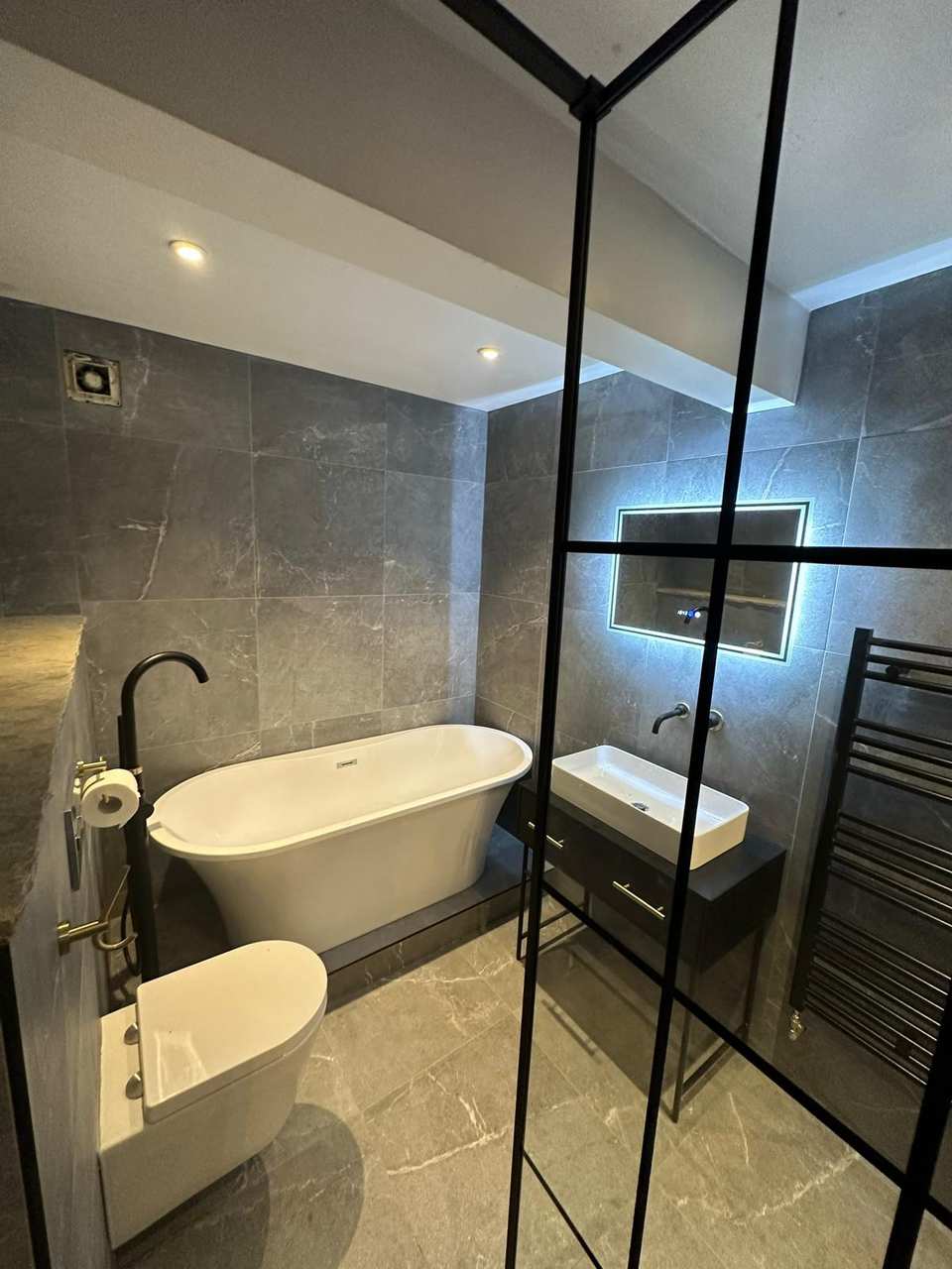Luxury finished bathroom with grey marble-effect tiles, freestanding bath, backlit mirror, and matte black fixtures.