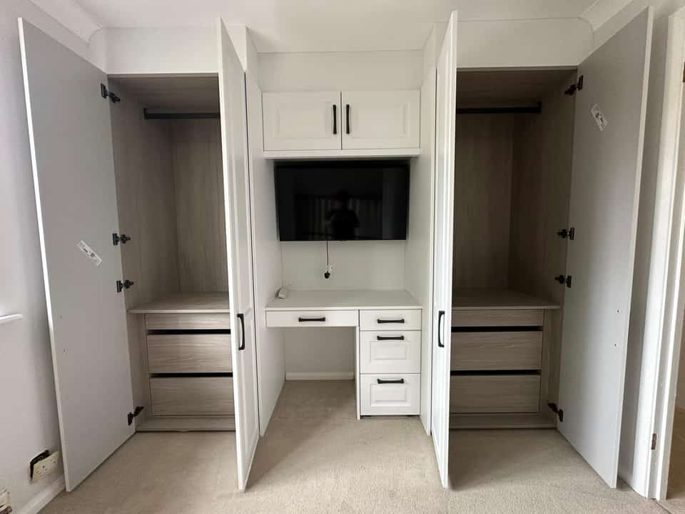 Custom white shaker bedroom storage unit with open oak-lined wardrobes, built-in desk, drawers, and wall-mounted television.