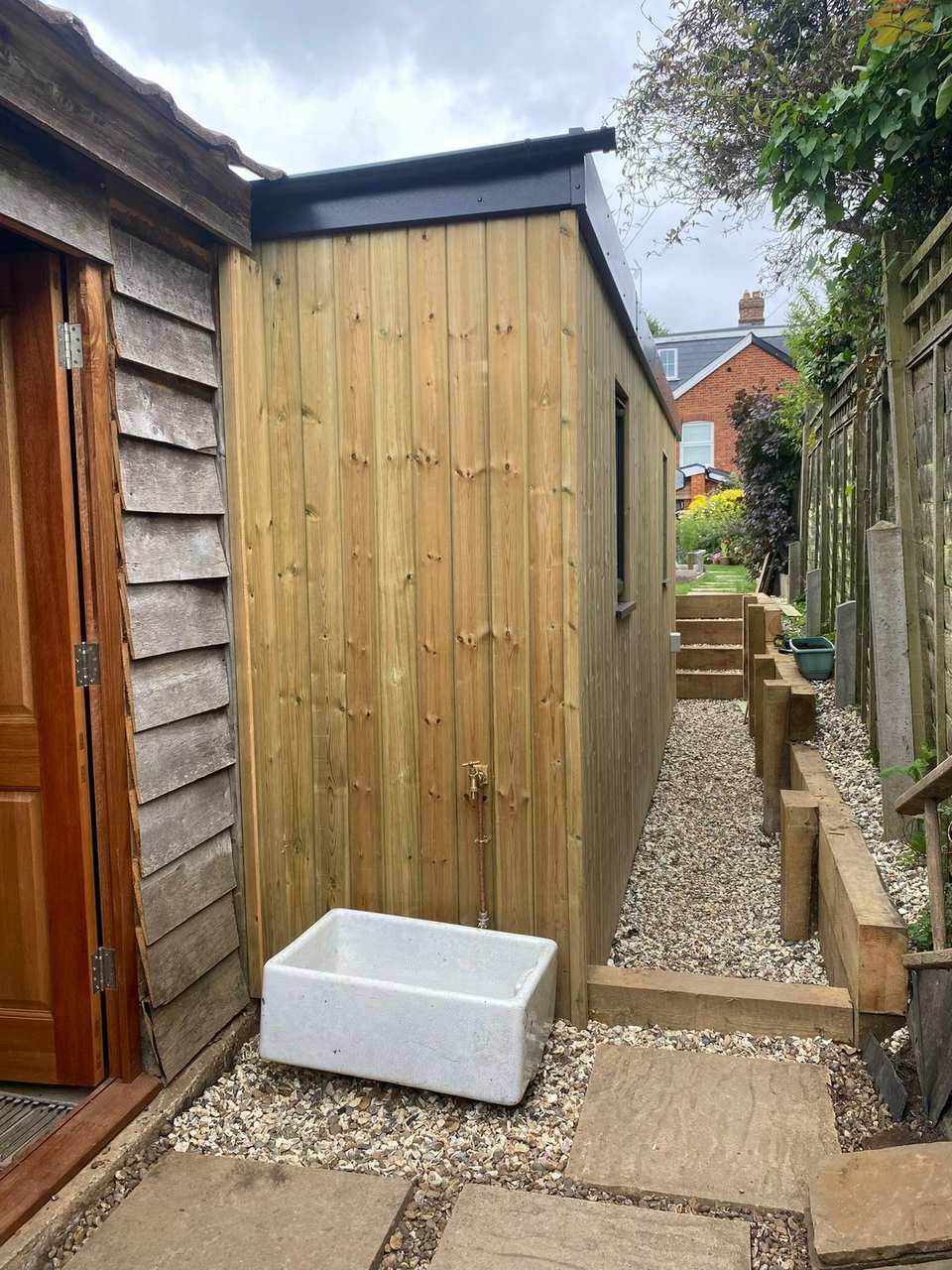 Finished vertical timber-clad garden room with flat roof, gravel path, sleeper retaining walls, and an outdoor Belfast sink.