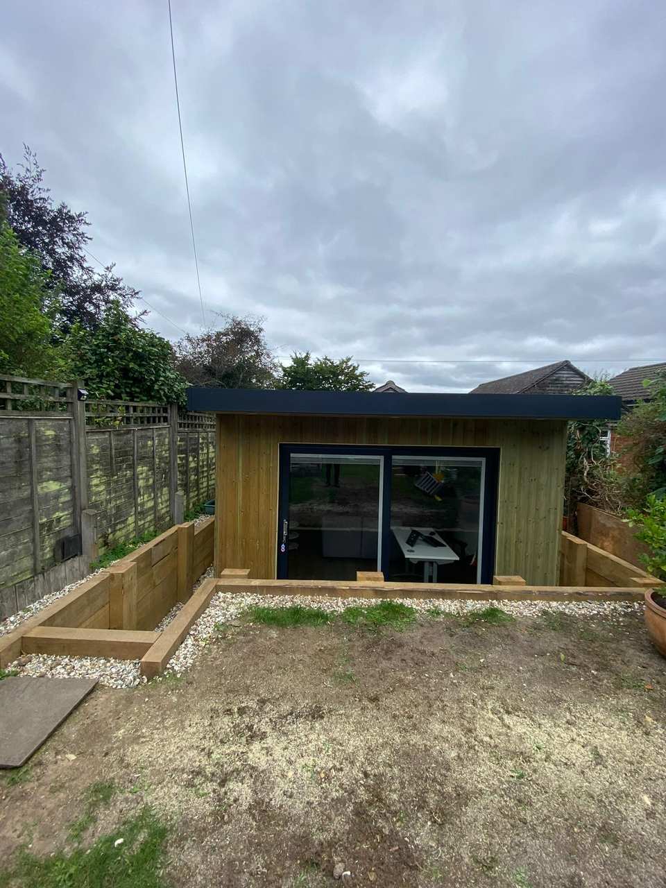 Completed timber-clad garden room with large sliding glass doors, set into a landscaped area with timber sleeper retaining walls.