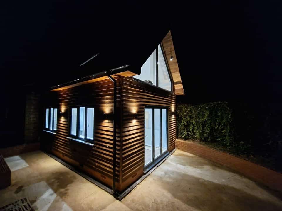 Timber-clad garden room at night with warm exterior wall lights, bifold doors, and triangular gable window.