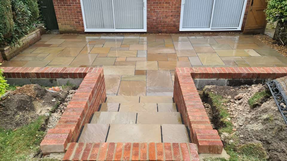 Newly laid multi-toned sandstone patio with red brick retaining walls and wide stone steps leading to a lower garden.