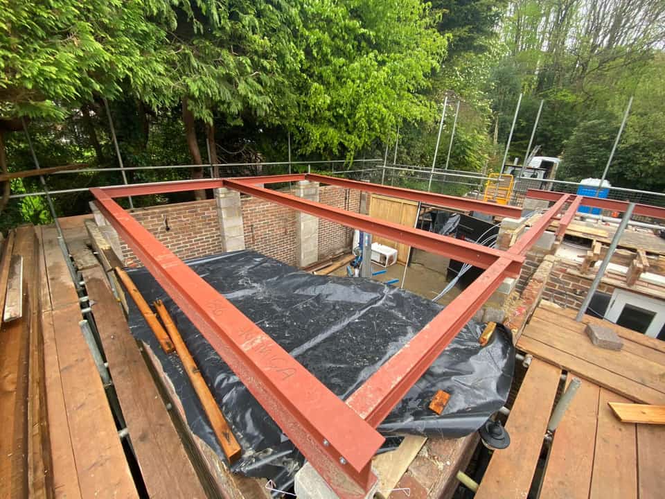 Red-painted steel RSJ beams installed on concrete padstones atop brick and block walls for a house extension, viewed from scaffolding.