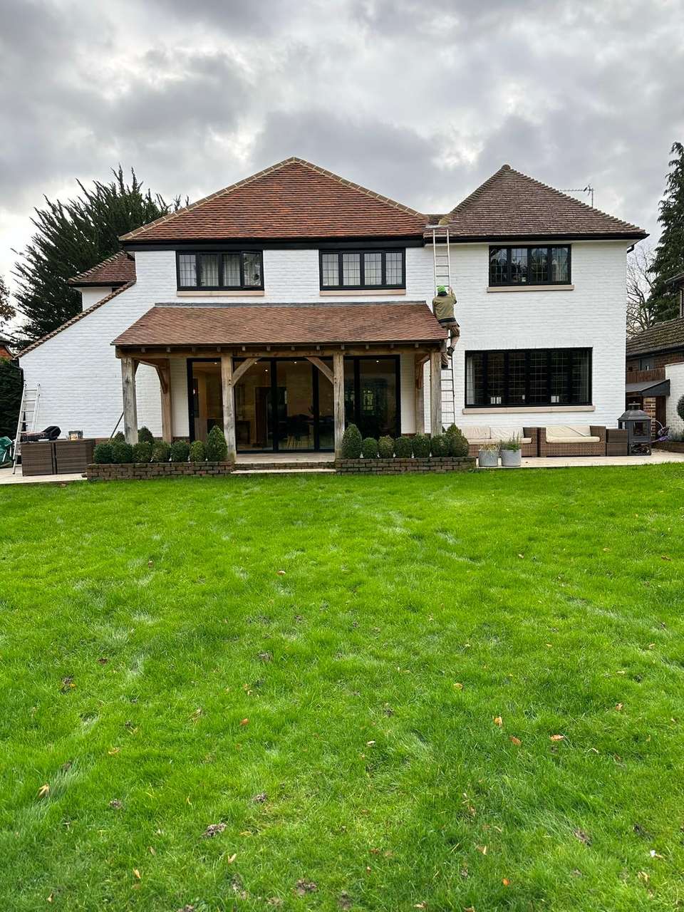 Wide garden view of a white brick house with a large oak-framed rear extension, bifold doors, lawn, and a worker on a ladder.