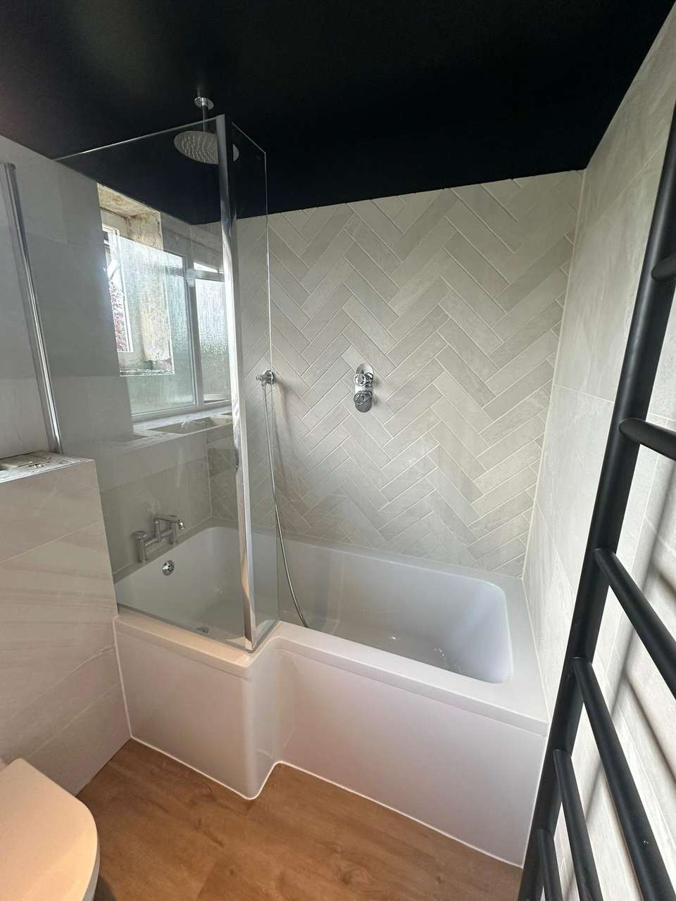 Modern bathroom with white L-shaped shower bath, herringbone wall tiles, chrome rainfall shower, and black ceiling.