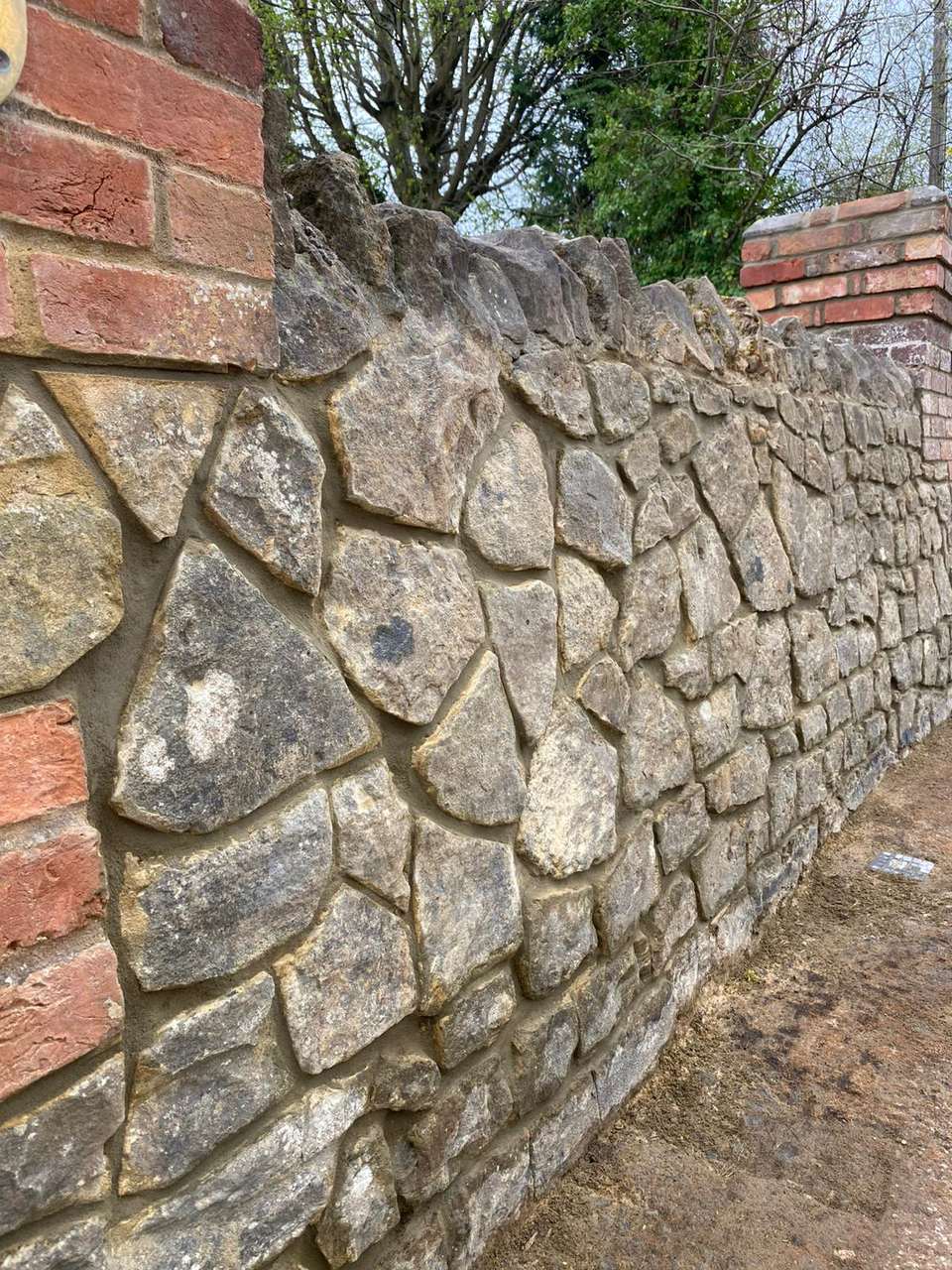 Close-up of a natural stone boundary wall with cock-and-hen coping, flanked by red brick pillars.