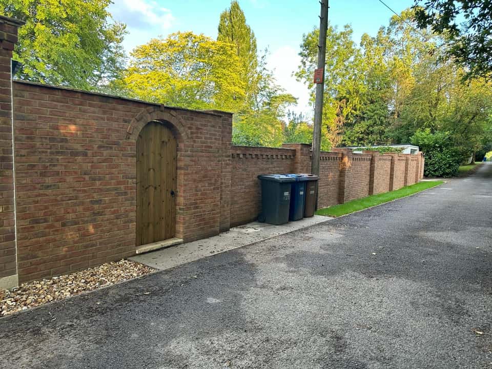 A long finished red brick boundary wall with decorative pillars and an arched wooden gate beside a driveway.