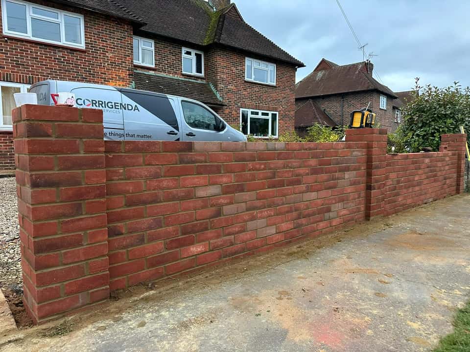 A newly built red brick boundary wall with square pillars and a laser level on top in front of a house.