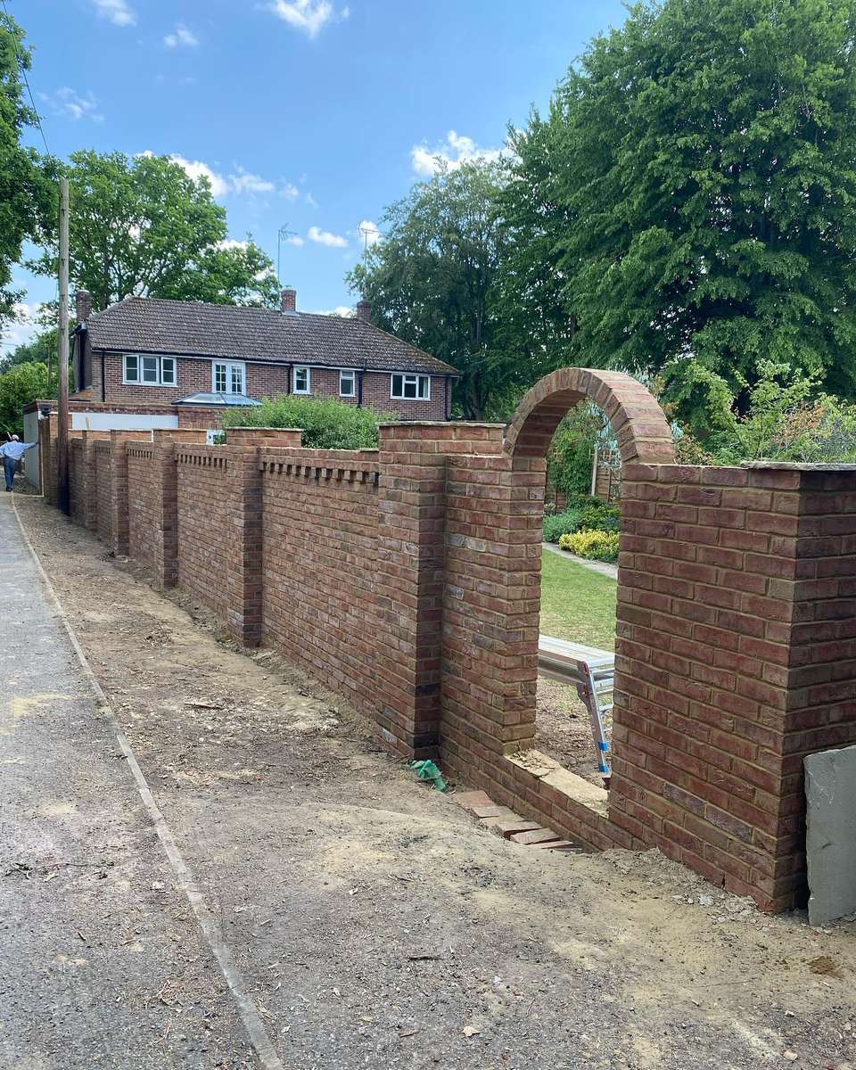 Long red brick boundary wall with dentil course, square piers, and a rounded archway opening beside a matching brick house.