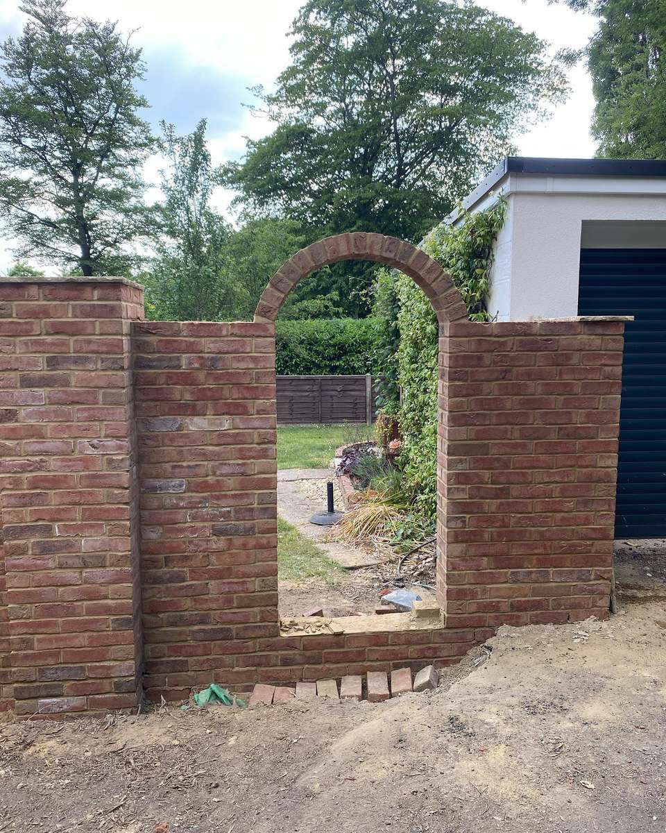 Red brick garden wall with a semicircular header-bond archway framing a view of a landscaped lawn and stone path.