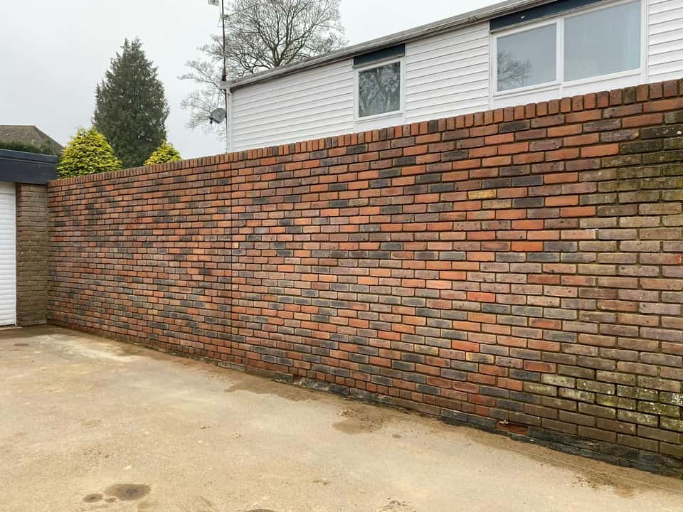 A long finished boundary wall built from multi-tonal red and grey bricks beside a concrete driveway.