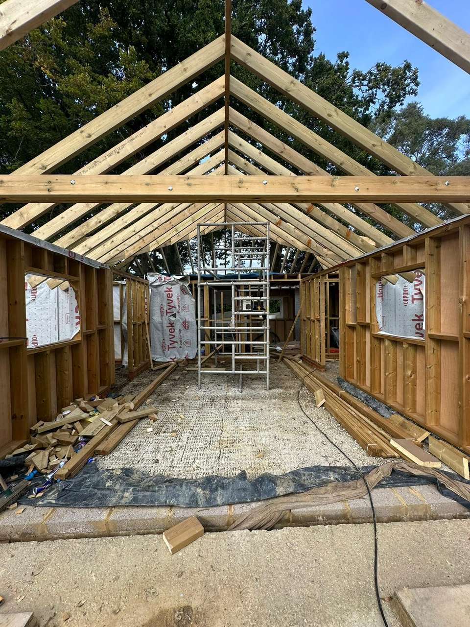 Interior view of a timber-framed garden room under construction with pitched rafters, Tyvek wrap, and scaffolding tower.