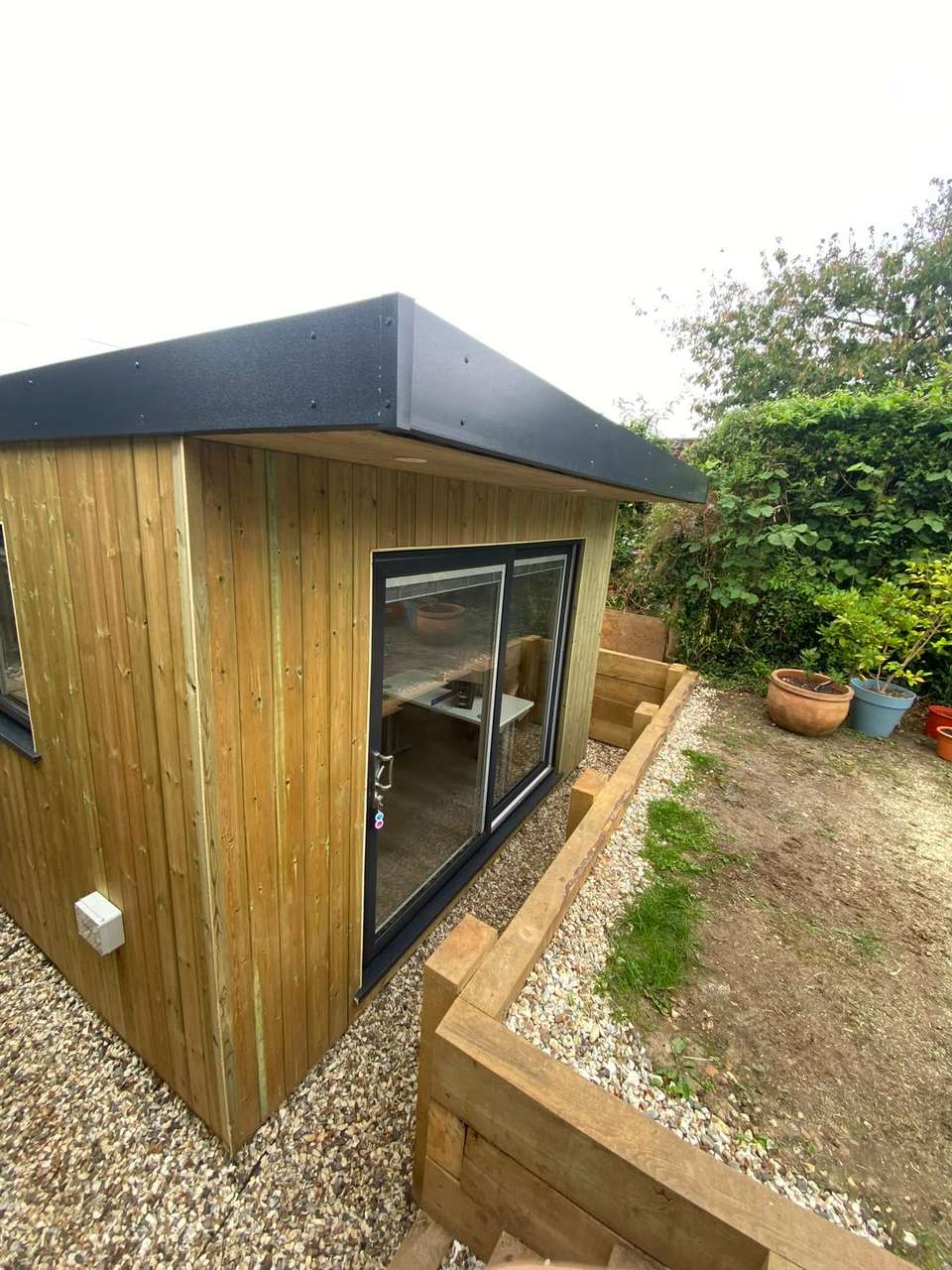 Completed timber-clad garden room with anthracite sliding glass doors and a flat roof fascia in a landscaped garden.