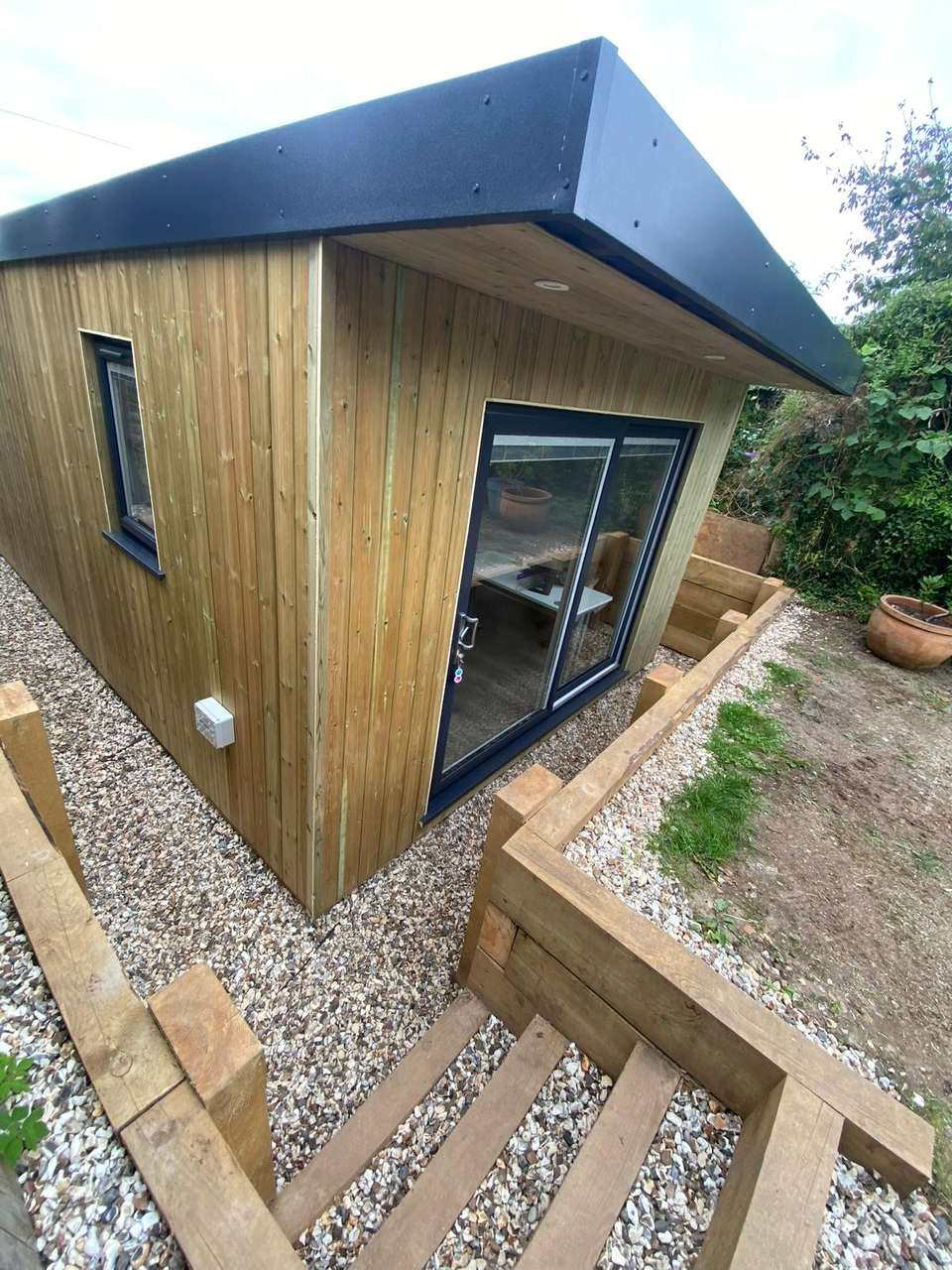 Completed timber-clad garden room with anthracite sliding doors and sleeper retaining steps.