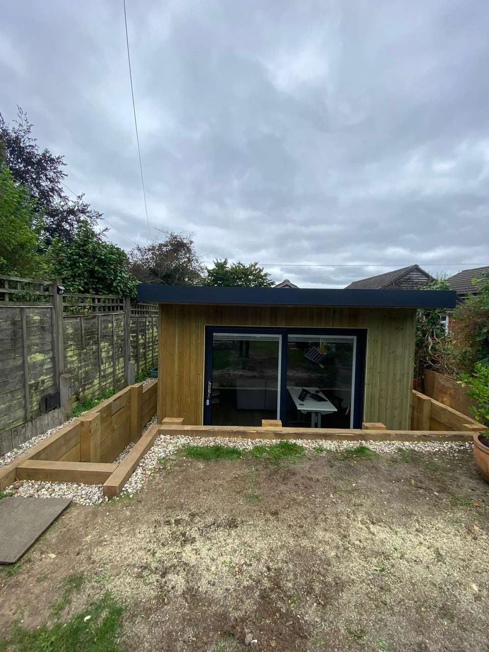 Completed timber-clad garden room with large sliding glass doors, set into a landscaped area with timber sleeper retaining walls.