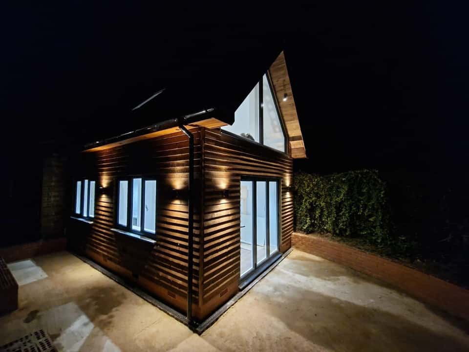 Timber-clad garden room at night with warm exterior wall lights, bifold doors, and triangular gable window.