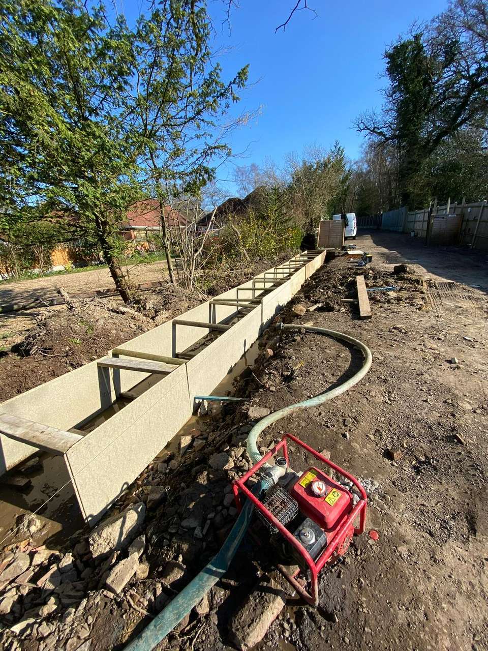 Long narrow construction trench lined with grey shuttering and wooden braces, with a red water pump clearing water from the excavation.