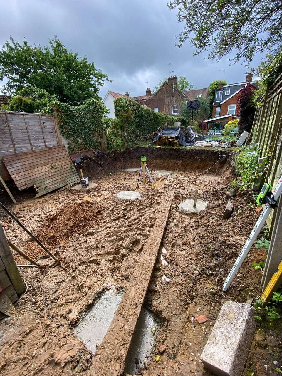 Excavated muddy back garden with circular concrete footings and a laser level on a tripod.