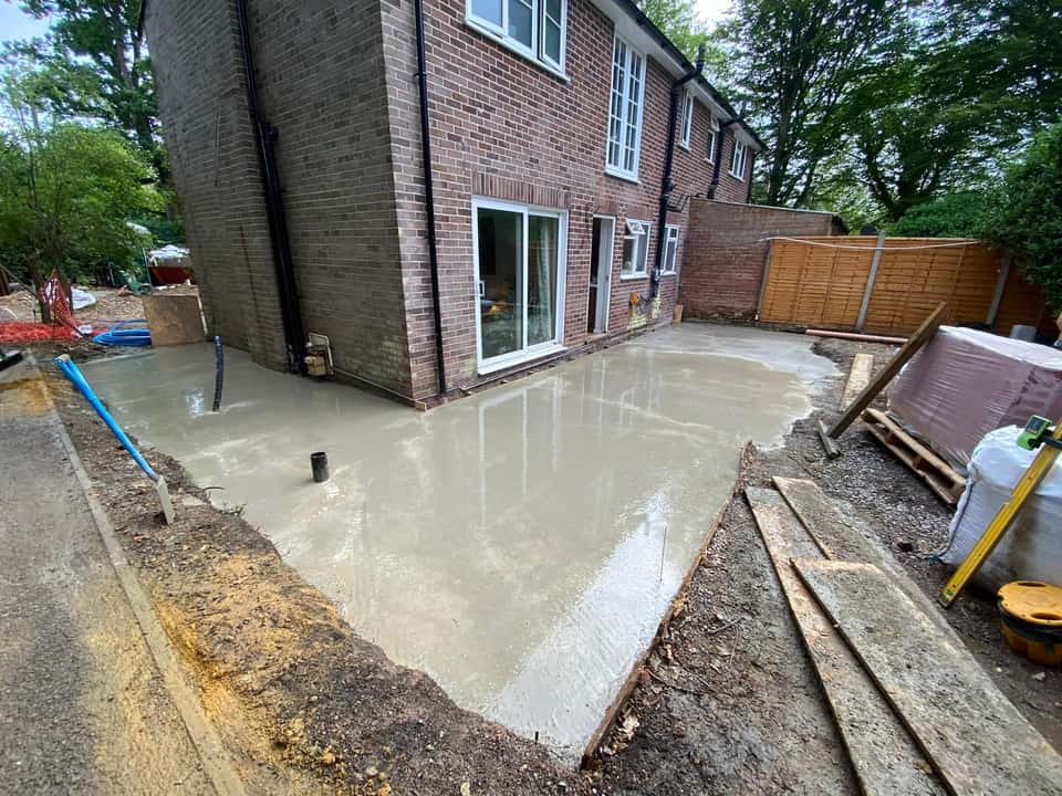 Fresh wet L-shaped concrete slab poured around the corner of a red brick house