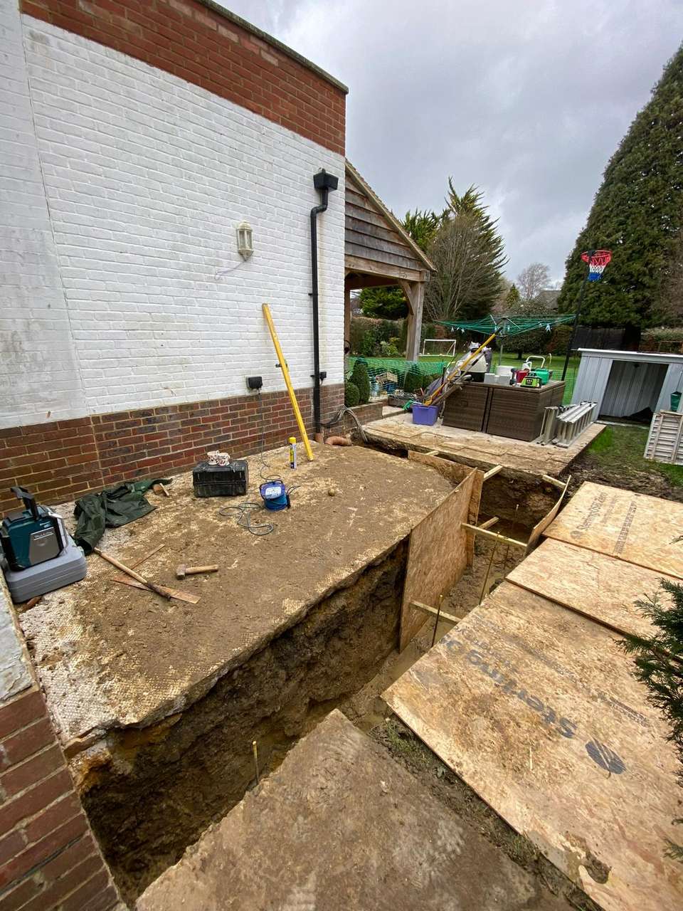 Deep foundation trenches excavated along the side of a house with OSB boards and construction tools on site.