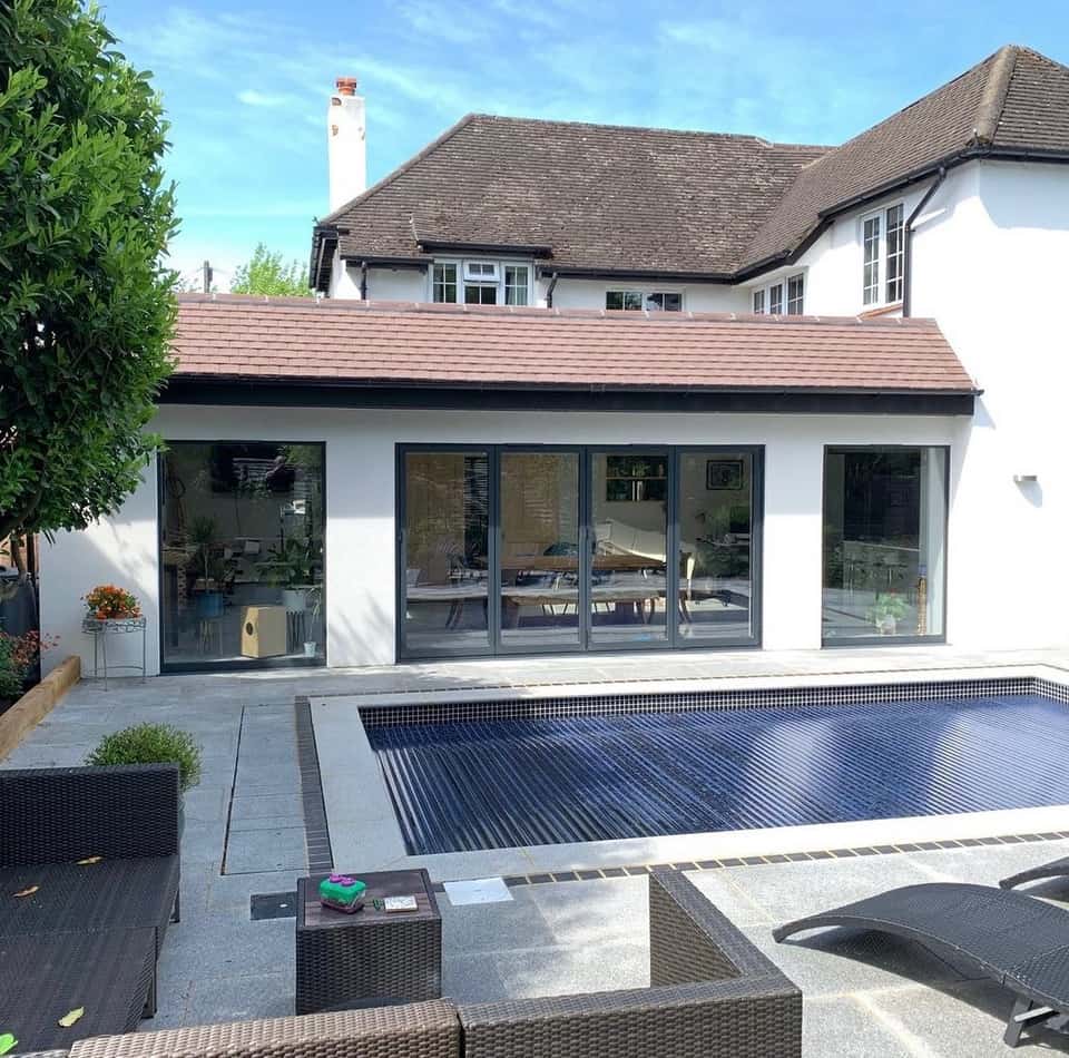 Completed white-rendered rear house extension with bifold doors, stone patio, swimming pool, and outdoor seating on a sunny day.