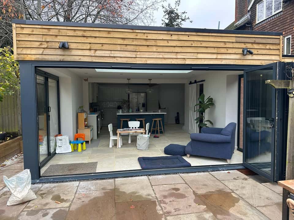 Completed rear extension with open anthracite bifold doors, timber cladding, and stone patio overlooking the garden.