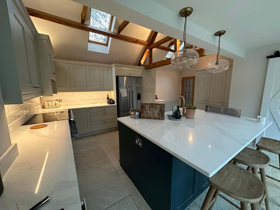 A bright modern shaker kitchen with a navy island, marble worktops, exposed timber beams, and skylights.