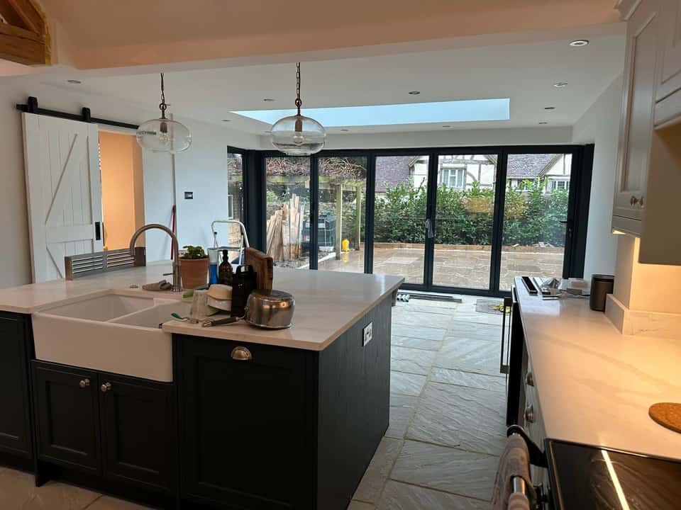 Finished open-plan kitchen with navy island, Belfast sink, bifold doors, roof lantern, and stone floor tiles.
