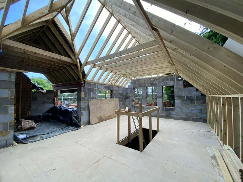 Interior loft conversion under construction showing exposed timber roof rafters, blockwork walls, and a staircase opening in the floor.