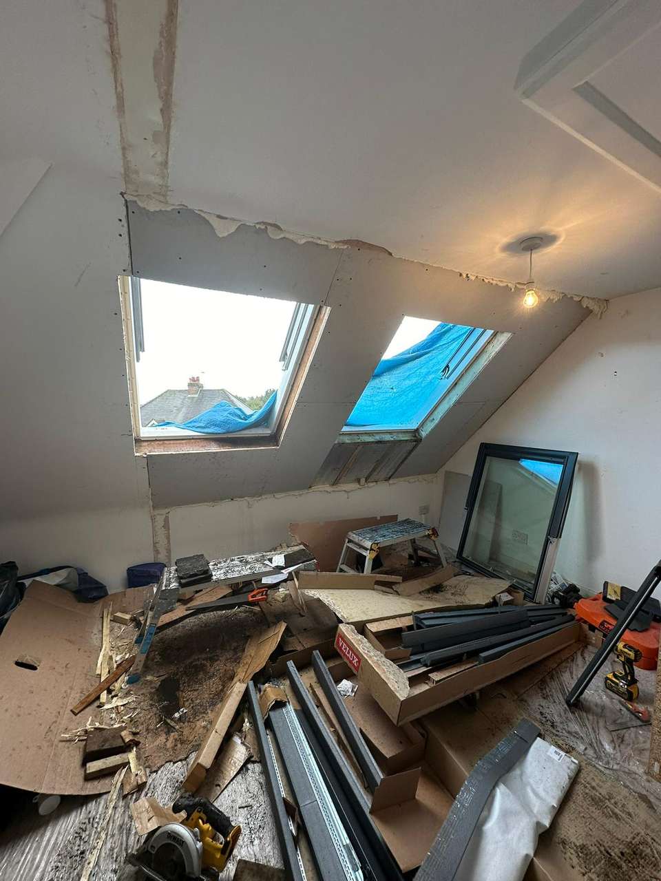 Loft conversion room with sloped ceiling, Velux skylight openings covered in blue tarp, and construction tools on site.