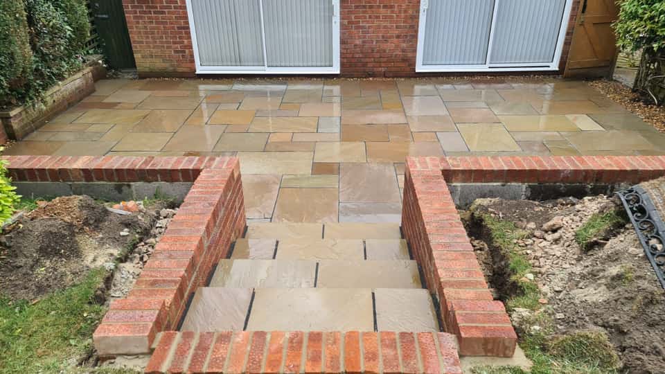 Newly laid multi-toned sandstone patio with red brick retaining walls and wide stone steps leading to a lower garden.
