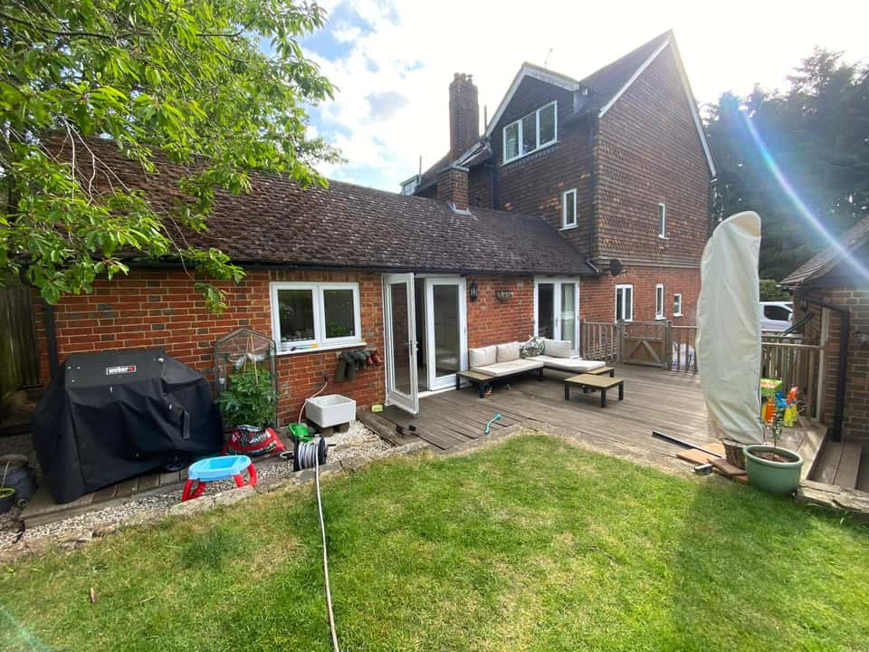 Rear garden with multi-level timber decking, outdoor seating, and a brick house beyond.