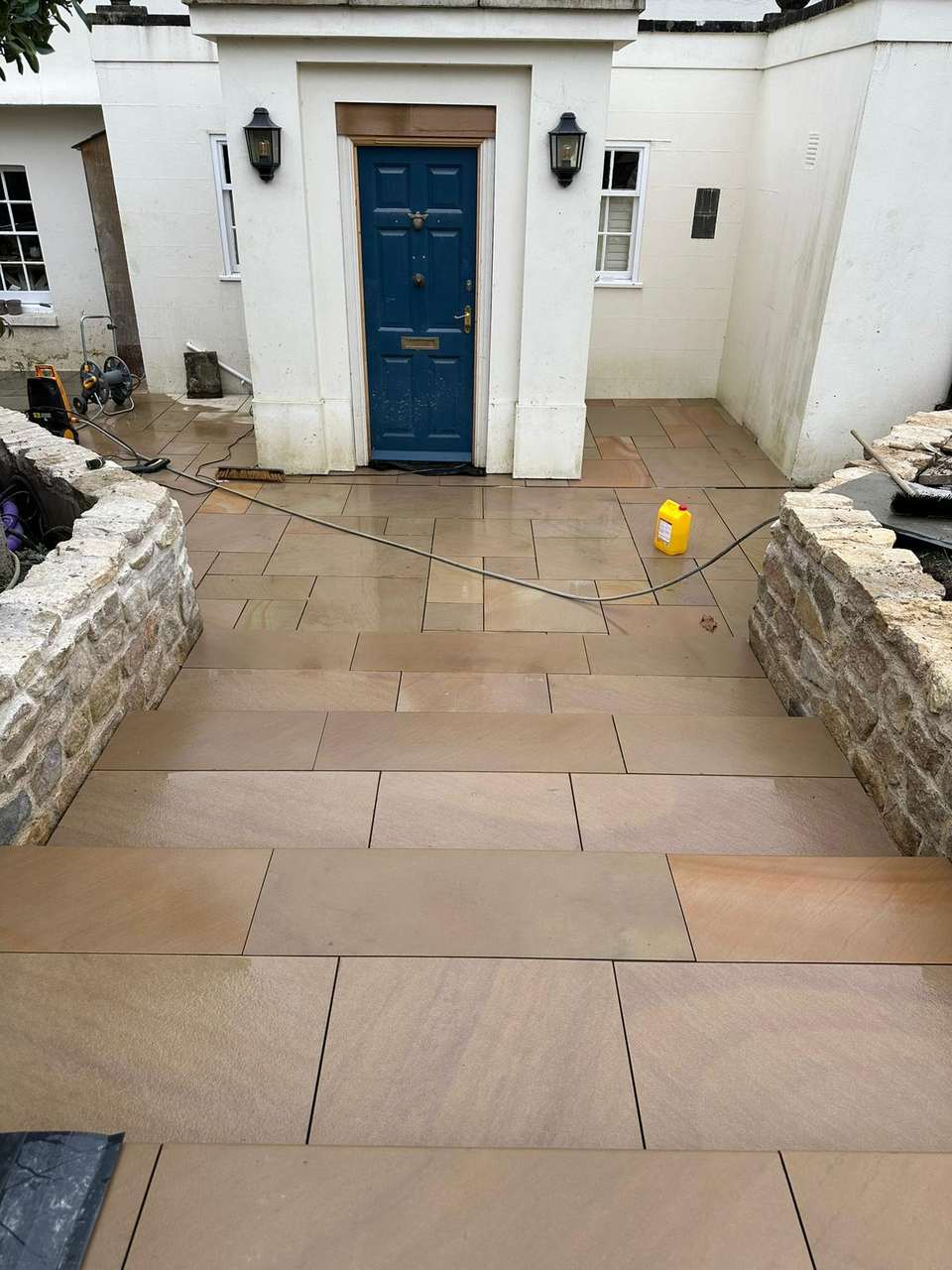 Newly laid buff sandstone patio and wide steps leading to a blue front door, bordered by rustic stone retaining walls.