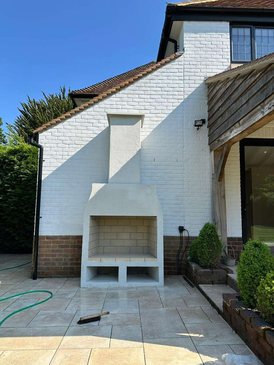 White rendered outdoor fireplace with brick-lined firebox and wood storage on a light stone patio.