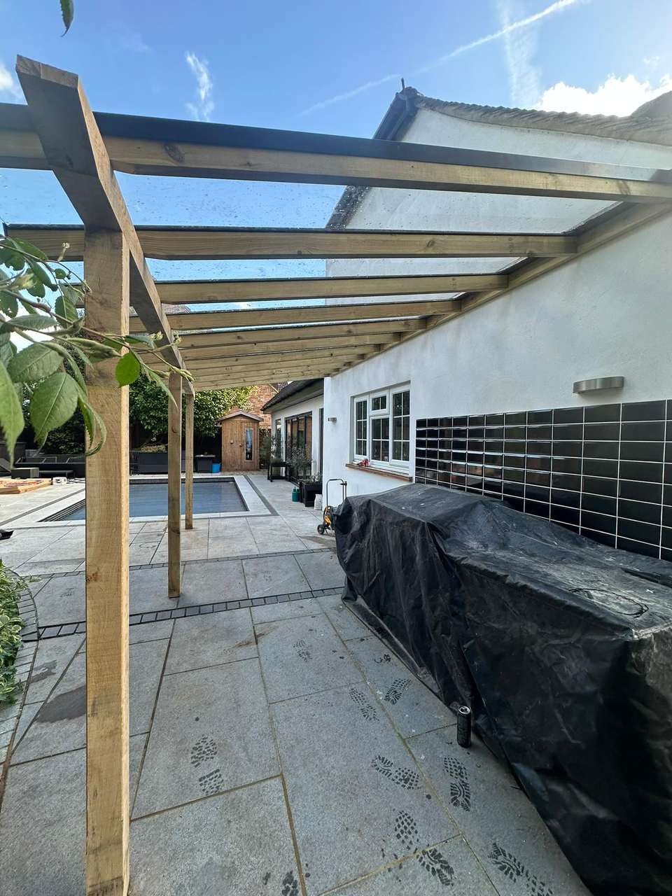 Large light-grey stone patio with a timber-framed pergola with glass roof attached to a white rendered house, with a pool beyond.