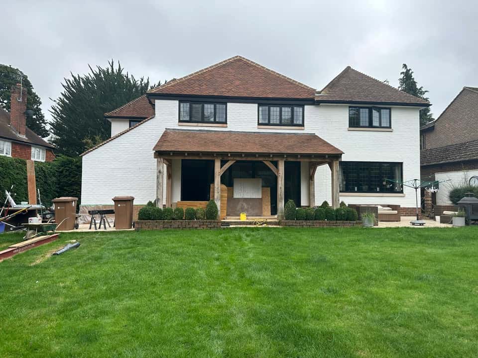 Wide rear view of a white brick house with a new oak timber porch, stone patio, and landscaped lawn.