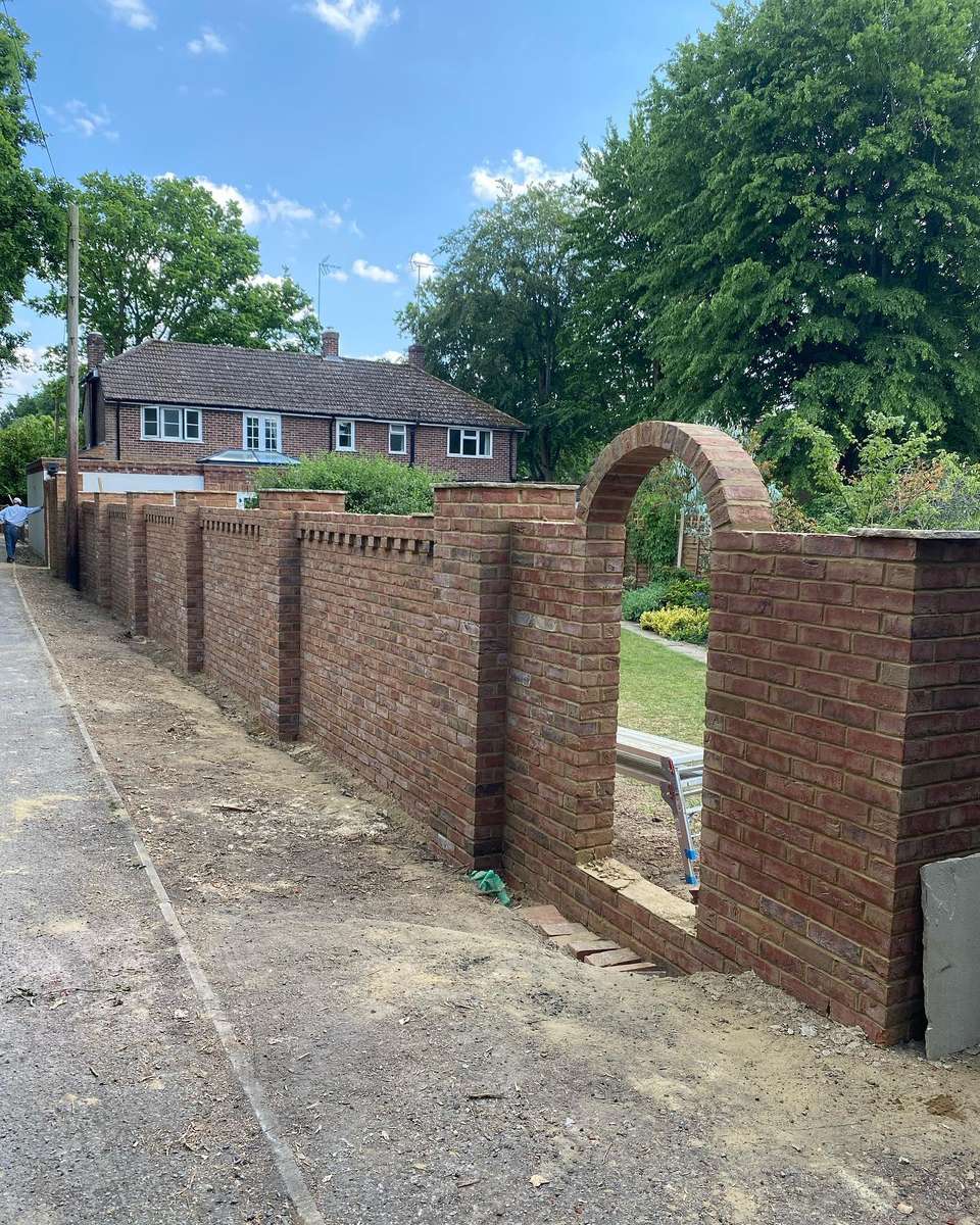 Long red brick boundary wall with dentil course, square piers, and a rounded archway opening beside a matching brick house.