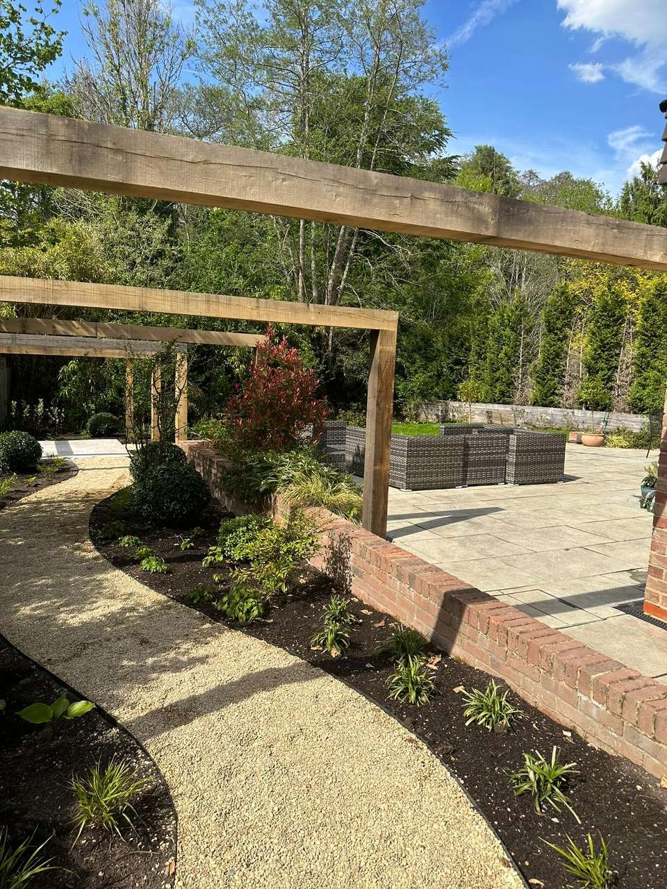 Completed luxury garden with large stone patio, rattan furniture, timber pergola, brick retaining wall, and gravel path.