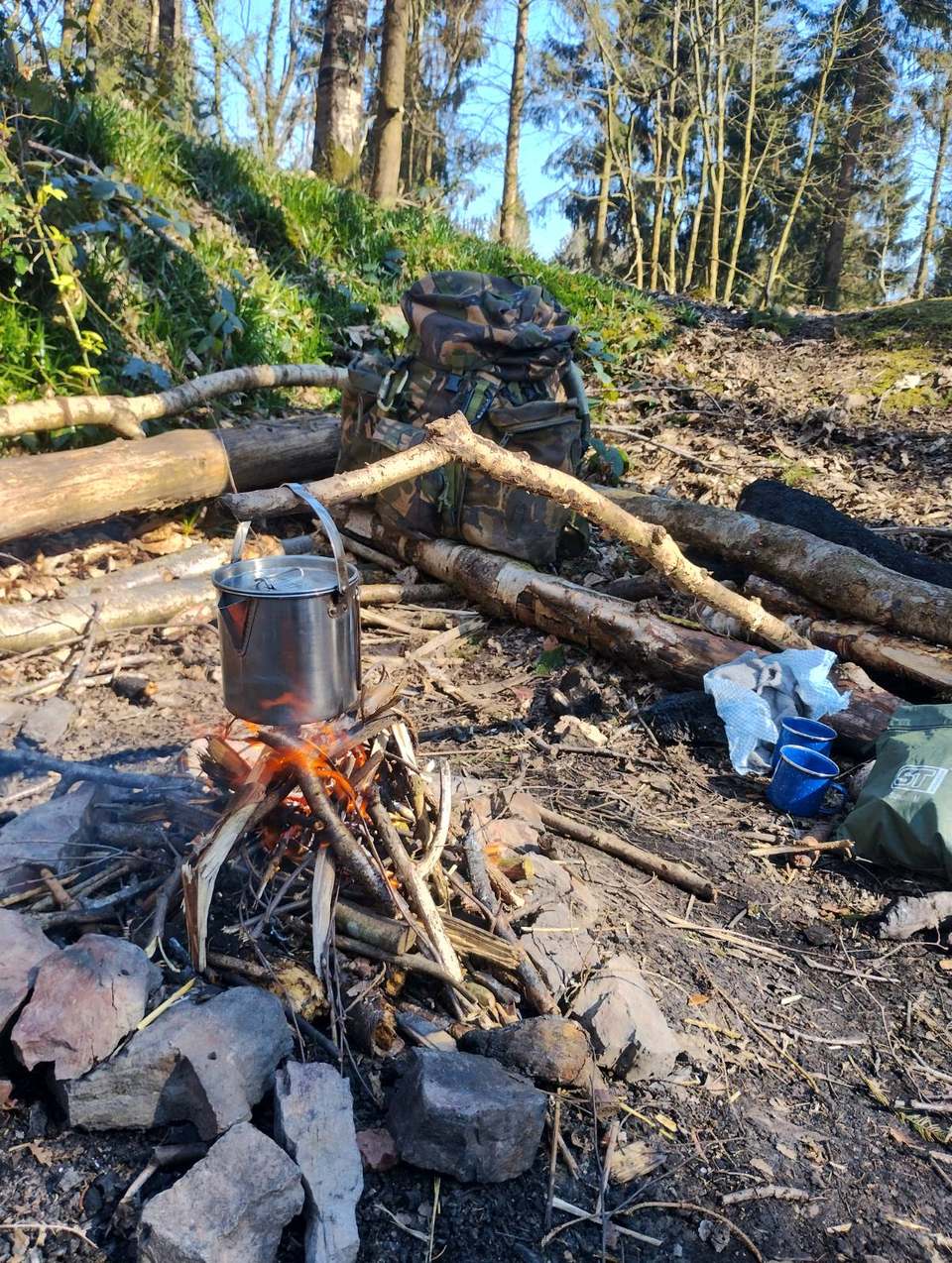 A small twig fire with billy-can and rucksack set up in sunlit woodland