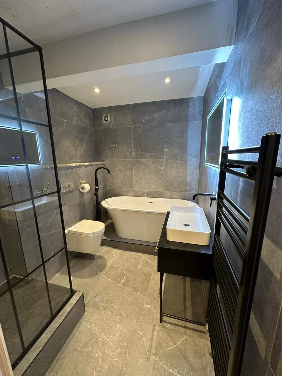Finished luxury bathroom with grey marble-effect tiles, freestanding bath, wall-hung toilet, and black fixtures.