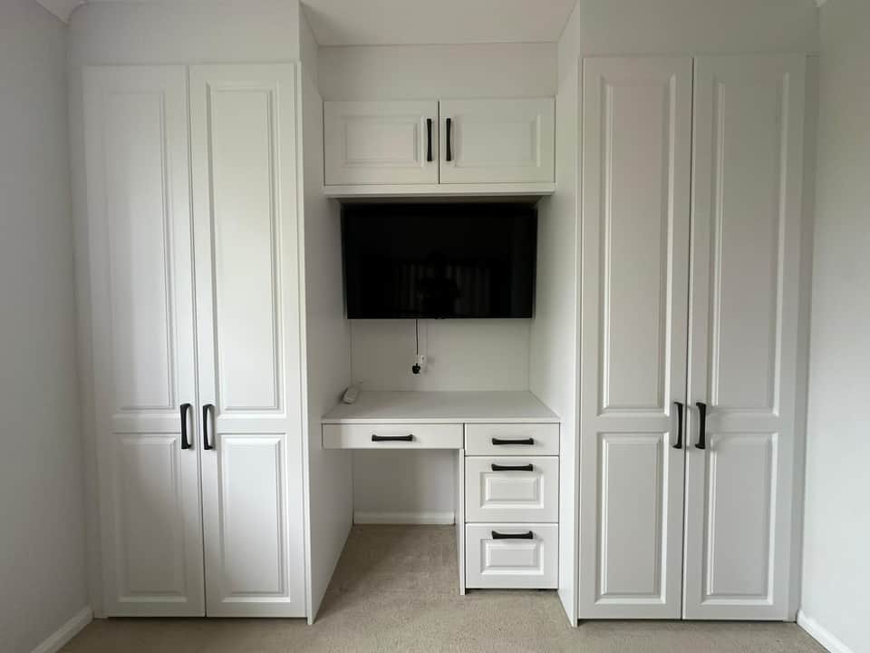 A floor-to-ceiling white shaker fitted wardrobe and desk unit with black handles and a wall-mounted TV.