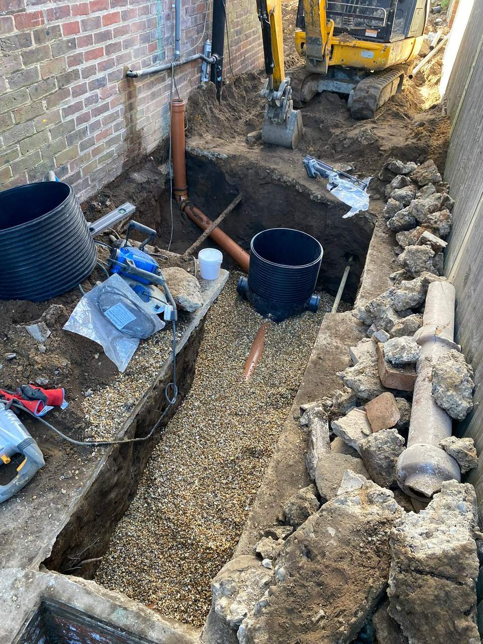 Excavated drainage trench with black inspection chamber, orange PVC pipes, and a mini excavator on site.