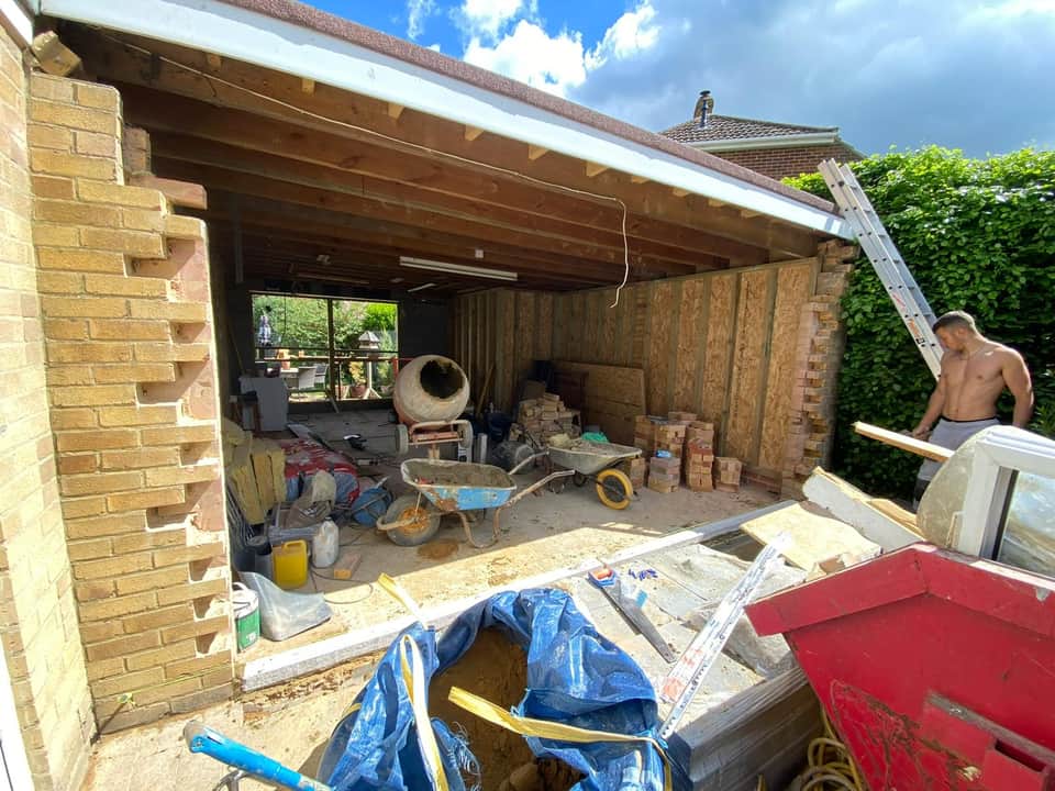 Garage conversion in progress with timber stud walls, exposed roof joists, cement mixer, and stacked bricks.