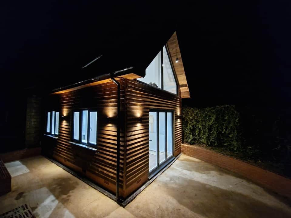 Timber-clad garden room at night with warm exterior wall lights, bifold doors, and triangular gable window.