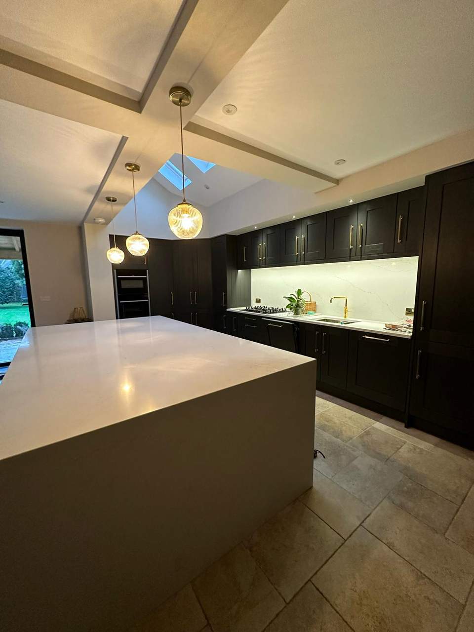 Luxury dark Shaker kitchen with white waterfall island, gold hardware, ribbed pendant lights, and a ceiling skylight.