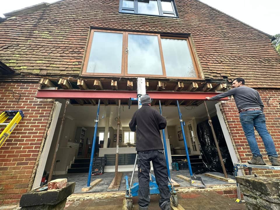 Two workers installing a red steel RSJ beam into a large opening in a red brick wall using a material lift.