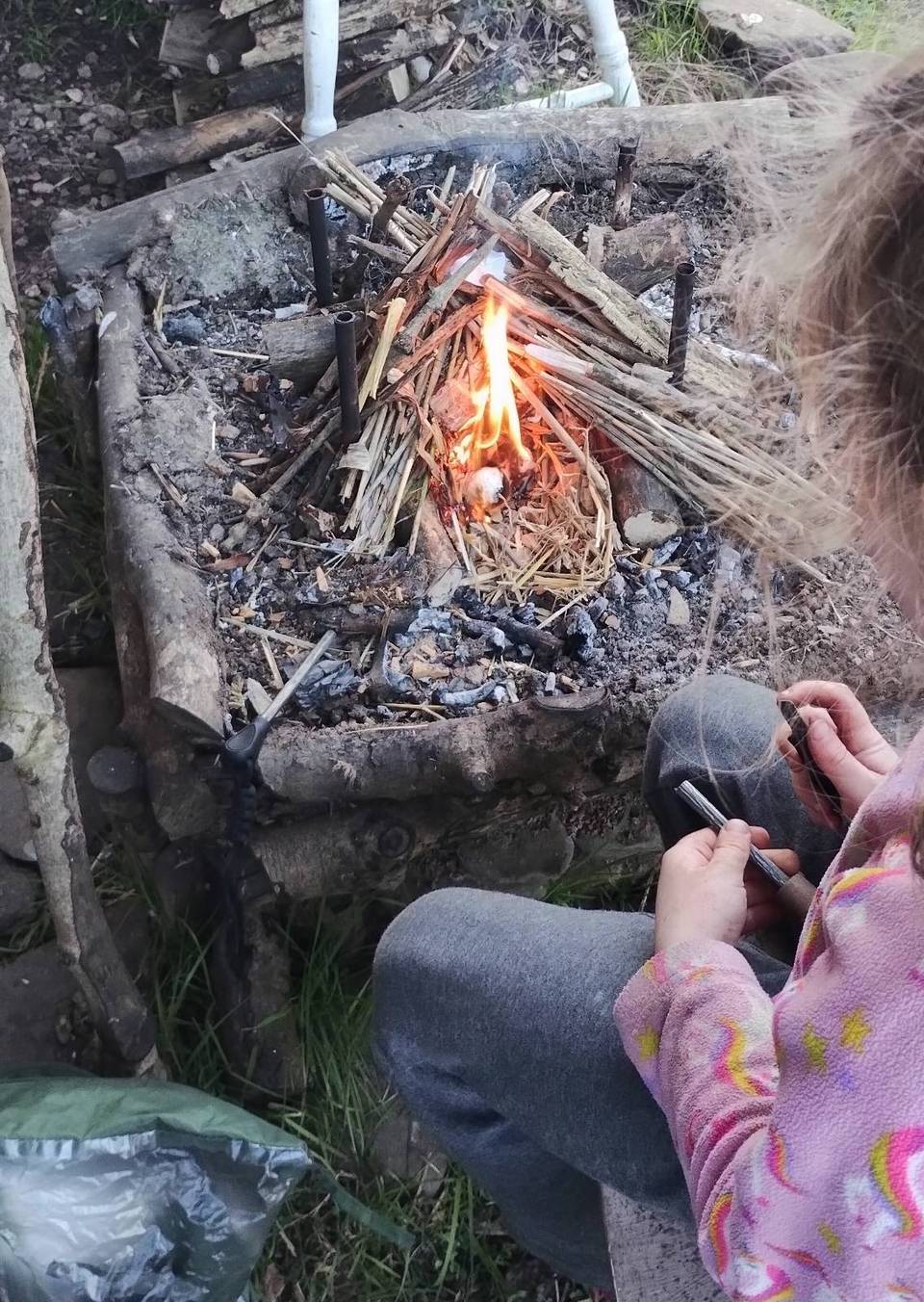 Child striking a ferro rod beside a teepee fire-lay in the woods