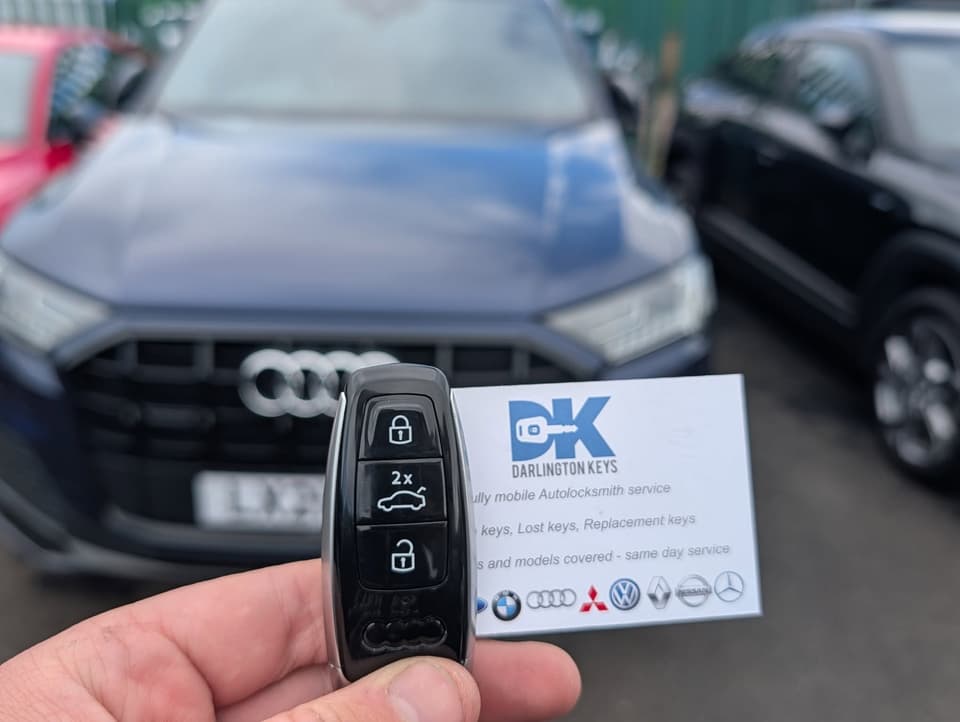 Hand holding a new Audi car key with Darlington Keys business card, Audi vehicles in background