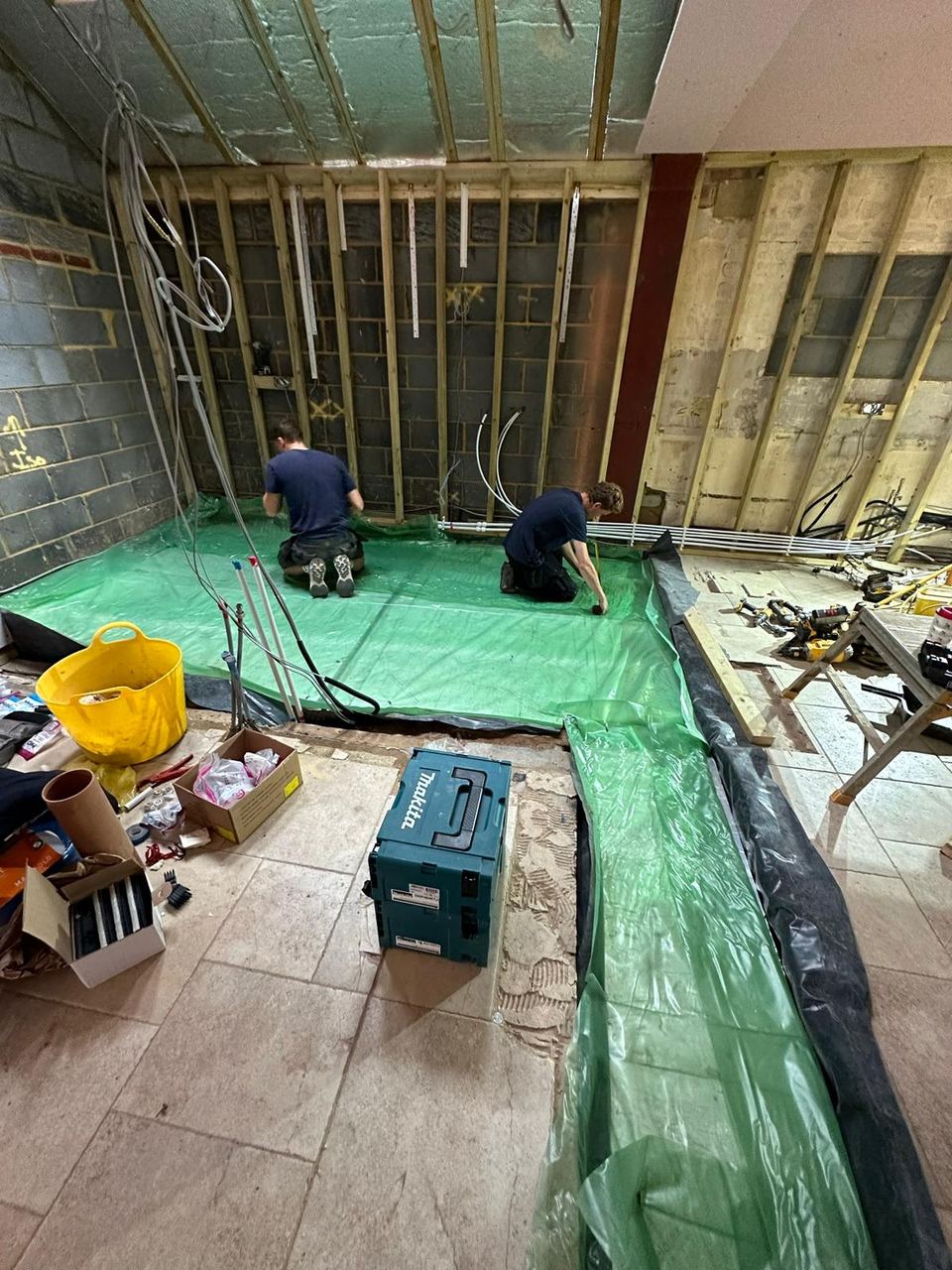 Two Woodhill Builders workers kneeling on green floor membrane inside a residential extension under construction.